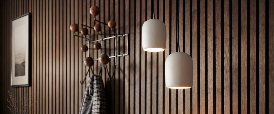 Tv&aring; vita f&ouml;nsterlampor h&auml;nger framf&ouml;r en m&ouml;rk ribbv&auml;gg och lyser upp hallen med kl&auml;dh&auml;ngare och konst. Den varma belysningen fr&aring;n Elbutik.se passar dekorativa entr&eacute;er och f&ouml;nstern&auml;ra ytor.