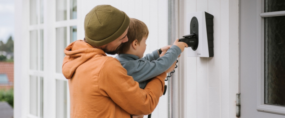 En man i orange tröja håller ett barn som kopplar in en laddkabel i en vit laddbox monterad på en husvägg. Elbutik.se erbjuder denna lösning för hem.