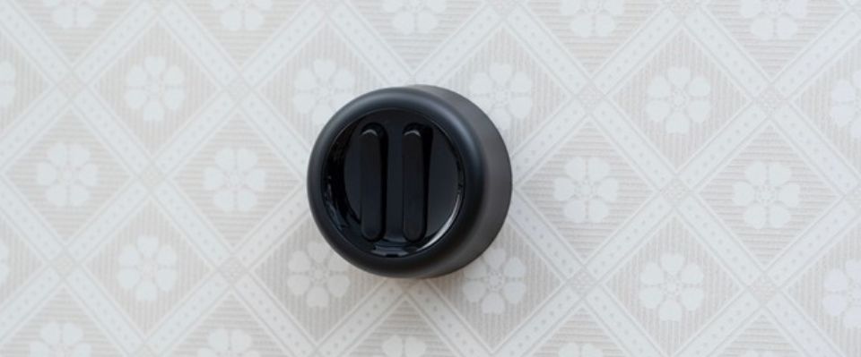 Svart lampknapp med tv&aring; smala reglage &auml;r monterad p&aring; en ljus, ornamentm&ouml;nstrad v&auml;gg och visar en stilren retrodetalj f&ouml;r belysningsstyrning, som Elbutik.se erbjuder f&ouml;r hem och milj&ouml;er med tydlig designk&auml;nsla.