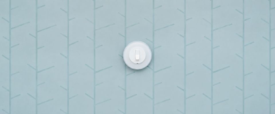 Vit lampknapp sitter centrerad p&aring; en bl&aring;gr&ouml;n tapet med grafiskt grenm&ouml;nster och anv&auml;nds f&ouml;r diskret styrning av belysning, en enkel och funktionell eldetalj som finns hos Elbutik.se.