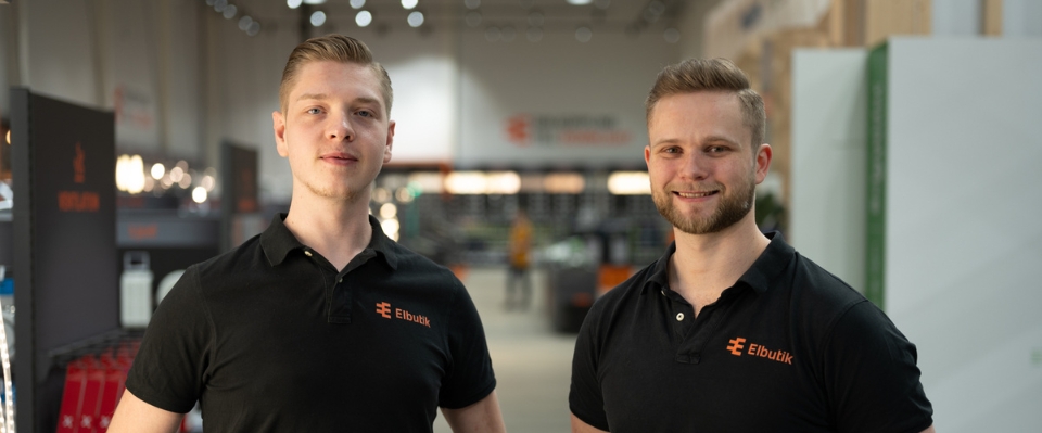 Två leende medarbetare från Elbutik.se står i en välorganiserad butiksmiljö. De har på sig svarta tröjor med Elbutik.se-logotypen, vilket representerar företagets vänliga kundservice och expertis inom elmaterial.